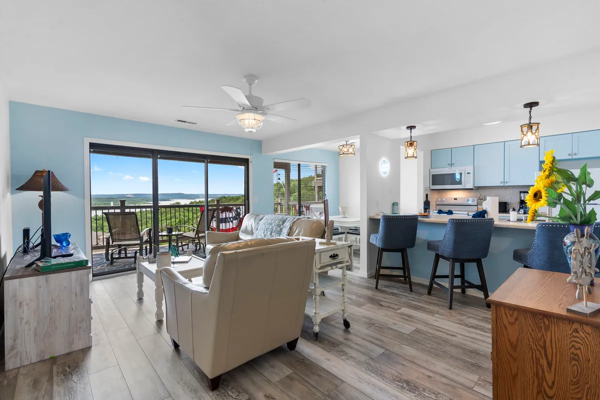 A bright, cheerful living area with modern kitchen amenities, comfortable seating by large scenic windows, and a decorative lamp creating a welcoming ambiance. The room is bathed in gentle natural light and offers views of the outdoors.