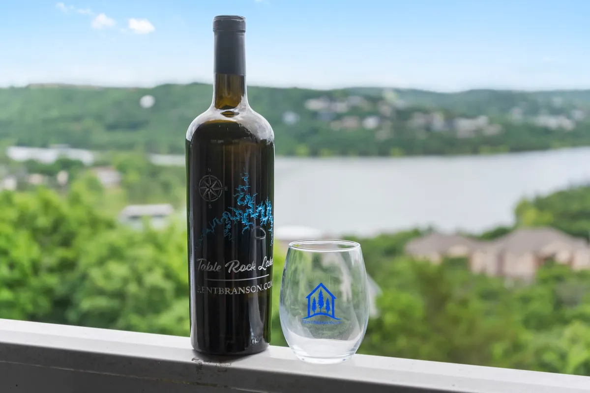 A close-up of a wine bottle and glass arranged on a table overlooking a peaceful lake. The view stretches over the water, framed by greenery, suggesting a relaxing moment enjoyed in the fresh air, possibly at sunset.