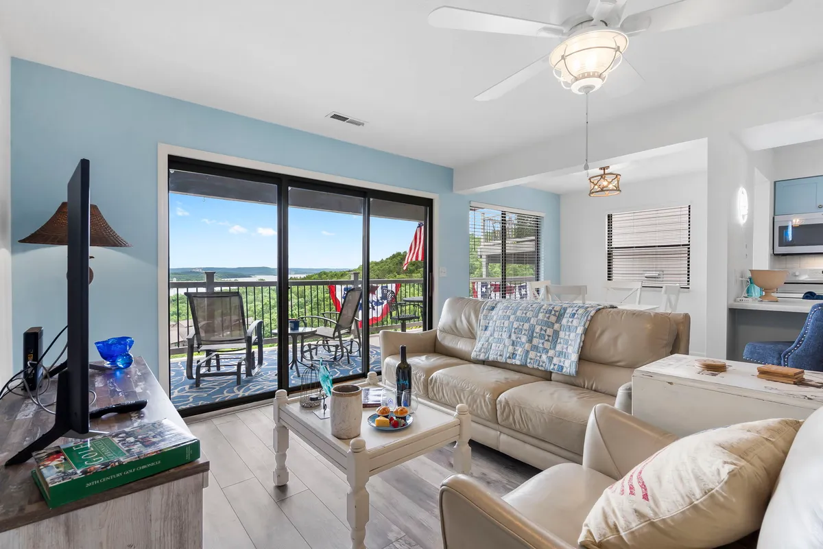 This image shows a comfortable beige sofa in a living area, positioned to face a large sliding glass door that opens to a scenic view of water and treetops. The wooden coffee table with decorative items adds warmth, and natural light pours in, inviting relaxation in a cozy, indoor-outdoor setting.