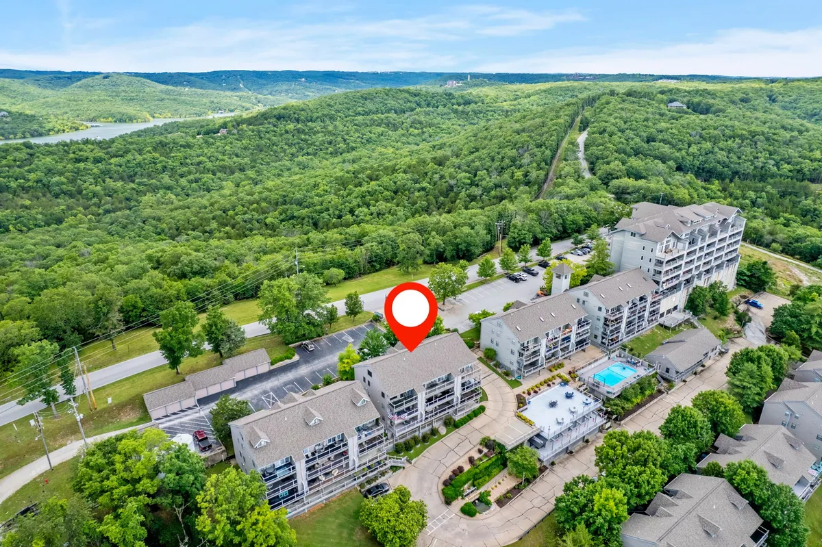 A wide view of the Silver Maple property's outdoor pool area, surrounded by green hills and several buildings. The pool is inviting, with lounge seating nearby, and the landscape in the background suggests a tranquil, semi-rural lakeside setting. The scene is sunlit and highlights the property's natural beauty and recreational amenities.