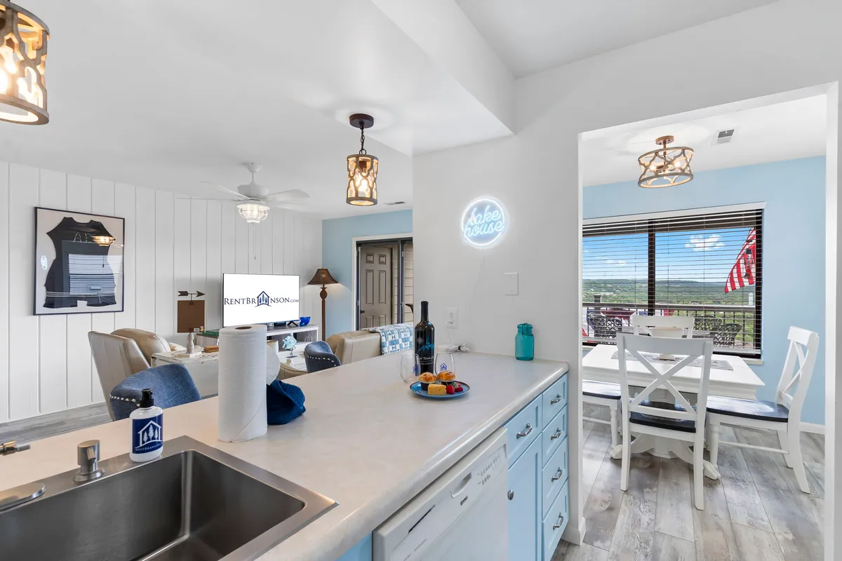 A spacious, open-concept living area featuring a kitchen island, adjacent dining area, and comfortable living room seating. Large windows flood the space with natural light, and a view outside hints at the relaxing lakeside setting.