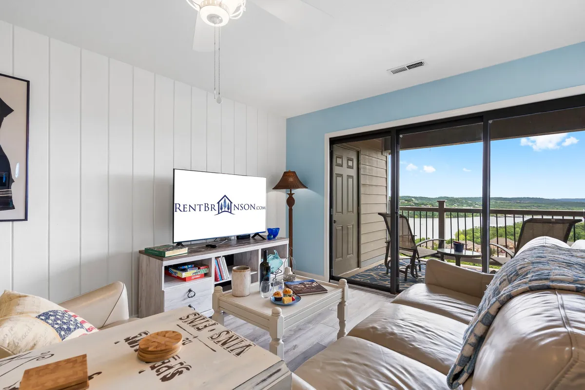 A cozy, inviting living area featuring a sofa, coffee table, large television, and a view out to the balcony overlooking the lake. The comfortable space is ideal for group gatherings and relaxation, with natural light streaming in from the balcony doors.