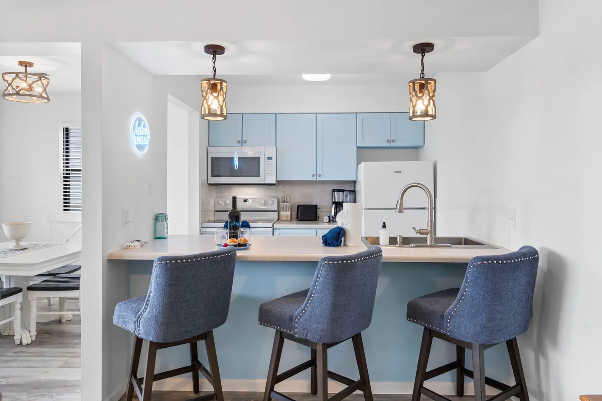 A vibrant kitchen scene highlights blue bar stools at a kitchen island, modern appliances, hanging light fixtures, and a lively, cheerful atmosphere. The image suggests spaces for both cooking and socializing.